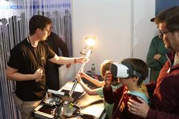 Ein Kind testet eine Virtual-Reality-Brille an einem HSD-Stand, während ein Mitarbeiter eine technische Versuchsanordnung mit Lichtquelle erläutert.